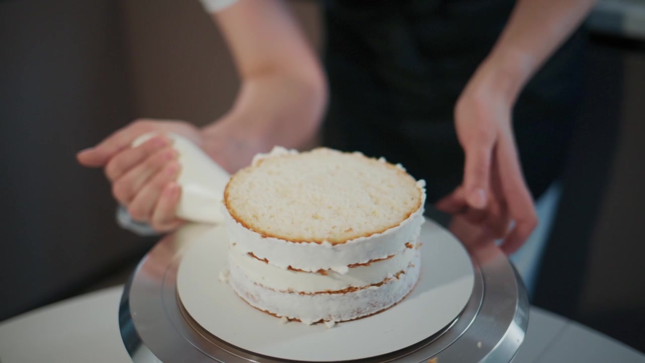 ケーキの組み立てのクローズアップ。ケーキの層にクリームが均一に塗られています。人物の手は安定しており、細部にまでこだわっています。レシピ動画やベーカリーのプロモーション、ケーキデコレーションの視覚的なガイドとして最適です。
