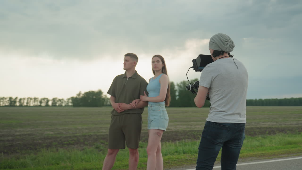 back view of videographer capturing couple standing on roadside field under cloudy sky with camera framing shot of couple gazing into distance amid rural landscape