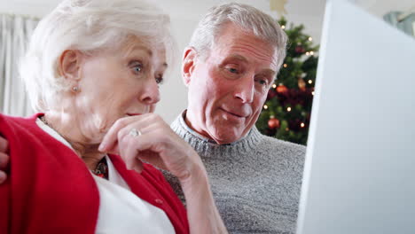 close up de una pareja de mayores sentada en el sofá usando una computadora portátil para comprar bienes o servicios en casa con el árbol de navidad en el fondo