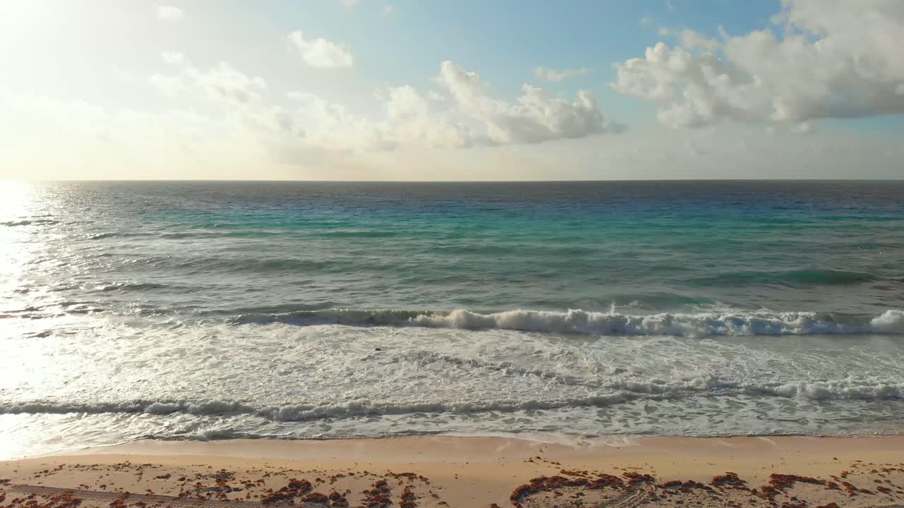Tropical sea waves crashing onto the white sandy shore next to seaweed, while golden sun rays shine through the luminous turquoise sea in Cancun, Mexico with white clouds in a blue sky - 4K drone