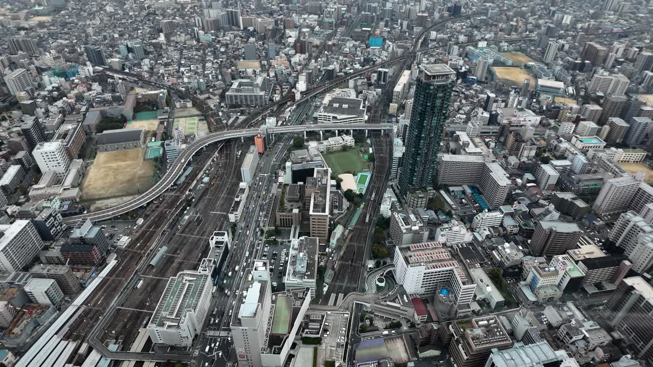 Looking down at the dense urban landscape of Osaka, Japan, featuring railway lines, skyscrapers, and a network of roads and buildings