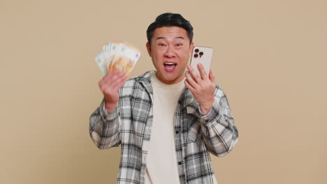 happy asian man use smartphone rejoicing game win receiving money euro cash celebrating success