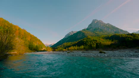 Mountain-river,-clear-blue-water-rushing-by-vibrant-colorful-autumn-trees-in-the-sun