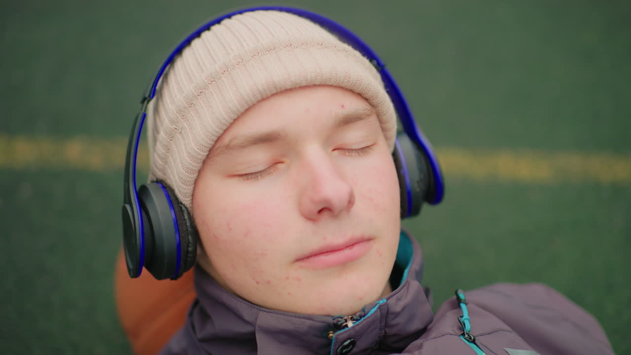 Giovane uomo sdraiato sull'erba, che ascolta musica con cuffie over-ear, indossa un berretto di lana e una giacca termica, occhi chiusi con espressione serena, linea di campo gialla sullo sfondo, ambiente esterno freddo