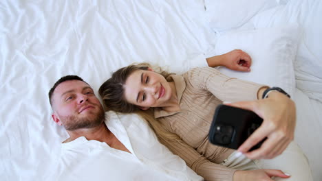 couple lies on the bed hugging. smiling blonde woman holding a phone taking selfie. top view.