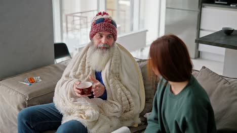 un anciano con un sombrero rojo y una manta blanca bebe té caliente de una taza marrón y se comunica con su hija adulta morena en una chaqueta verde mientras está sentado en el sofá en un apartamento moderno