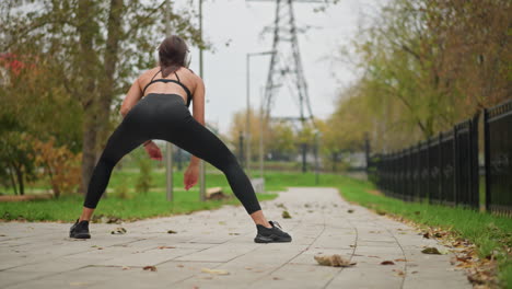 Back view of woman stretching her legs while listening to music with headphones, outdoor exercise in park, iron fence and trees visible in background, fitness and health routine