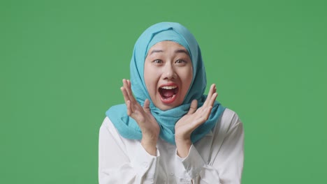 녹색 스크린 배경 스튜디오에 서있는 동안 미소 짓고 와우라고 말하는 아시아 무슬림 여성의 클로즈업