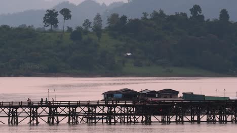 Mon-Bridge-and-a-longboat-followed-speeding-behind-the-structure-while-people-are-walking-on-the-bridge,-silhouetting-as-it-was-getting-dark,-in-slow-motion