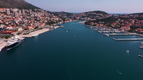 Amazing-reveal-shot-flying-backwards-from-harbour-to-sea-in-Dubrovnik,-Croatia-with-mountains,-boats-and-crystal-clear-water-in-4k-panning-down