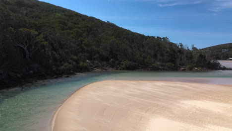 Revealing-drone-shot-of-the-Korogoro-Creek-and-coastal-mountains-with-wind-blowing-sand-across-a-sand-bar-at-Hat-Head-New-South-Wales,-Australia