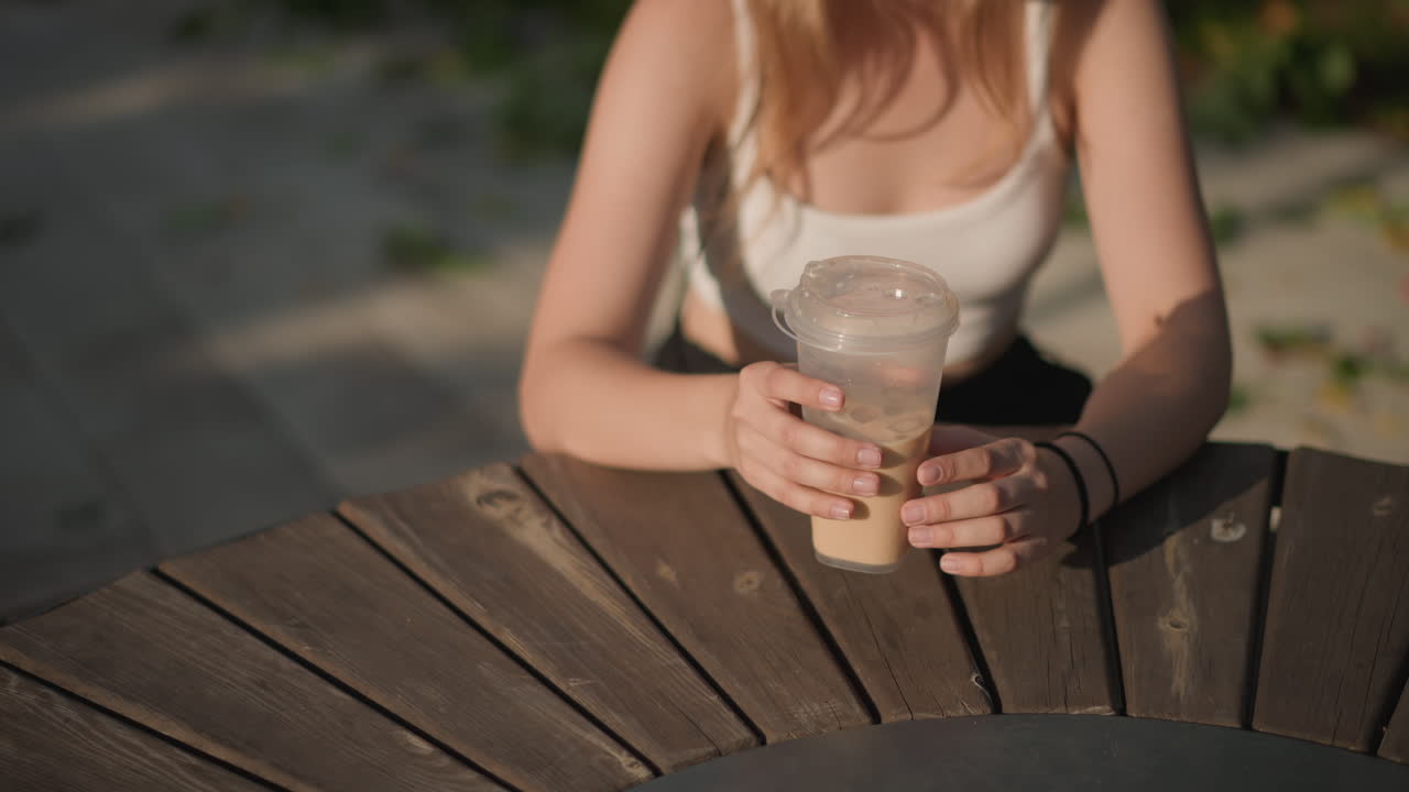 tavolo all'aperto illuminato dal sole con caffè freddo, giovane donna seduta a un tavolo rotondo di legno che tiene in mano un bicchiere di plastica, dita attorno al coperchio, luce dorata calda e ombre delicate sul marciapiede, canotta casual