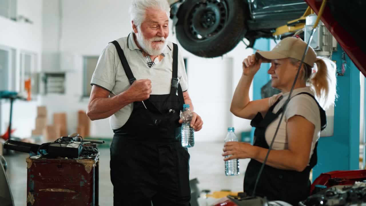 Mechanic workers rest while talking during a break in a workshop for repairing damaged cars. Old mechanic and an adult woman drink water during a break at work