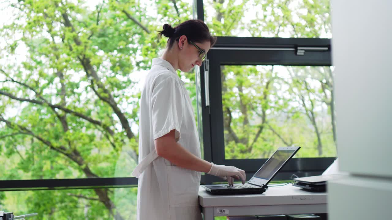 Female worker in laboratory checking test tube with blood sample and typing laptop keyboard, side middle shot, green natural background