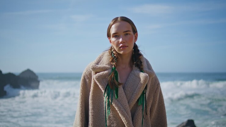 Ragazza alla moda in posa sull'oceano con un cappotto, ritratto. Donna attraente con le trecce sulla riva.