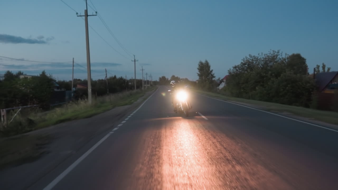 Biker rides motorcycle confidently along evening road as headlight shines brightly, reflecting on asphalt with power lines, houses, and roadside trees visible under fading sky during night approach