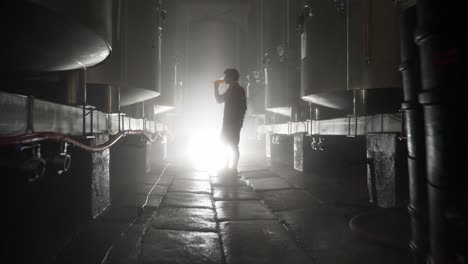 a guy drinking beer from a tank in brewery in glass with a contrast light behind him