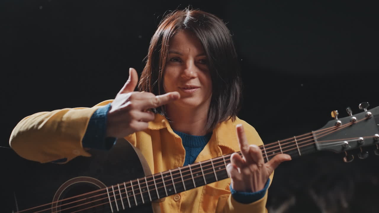 Ung musiker, der klimprer på akustisk guitar, nærbillede i studie med gul jakke, stemmer strenge, legende fingerbevægelser, udtryksfuldt smil, intim aftenoptræden med blødt spotlys og et oprigtigt udtryk.