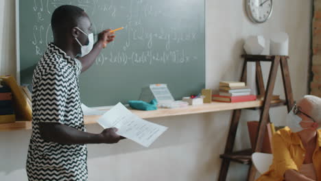 Afro-American-Student-in-Mask-Explaining-Formulas-on-Chalkboard