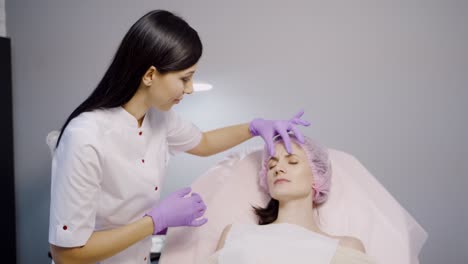 a beautiful cosmetologist advises a client who lays on a chair and examines her facial wrinkles in the aesthetic beauty salon on a gray background.