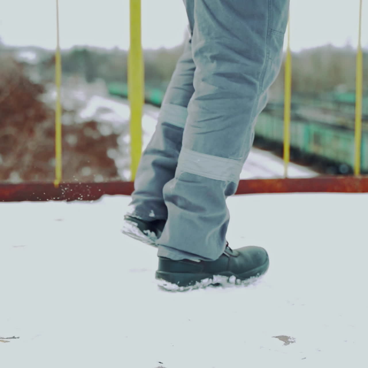 A man in a gray uniform climbs up the rusty stairs to the bridge and walks on it above the railway. Close-up on legs. Back view. Blurred background.