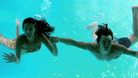 happy couple holding hands underwater in swimming pool and waving