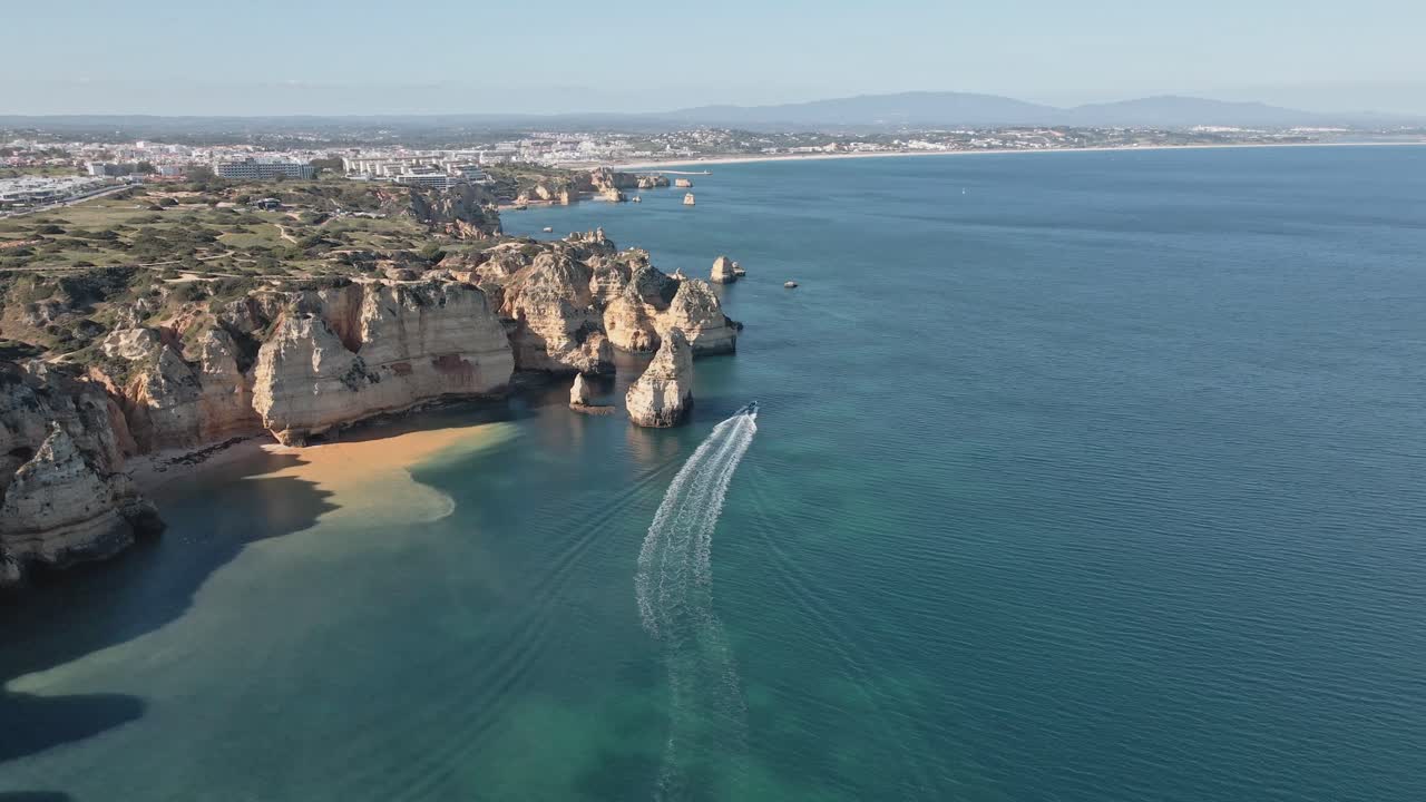 Aerial drone advances above the pristine cliffs and shallow waters of Lagos, Portugal, matching the forward motion of a boat speeding along the turquoise sea