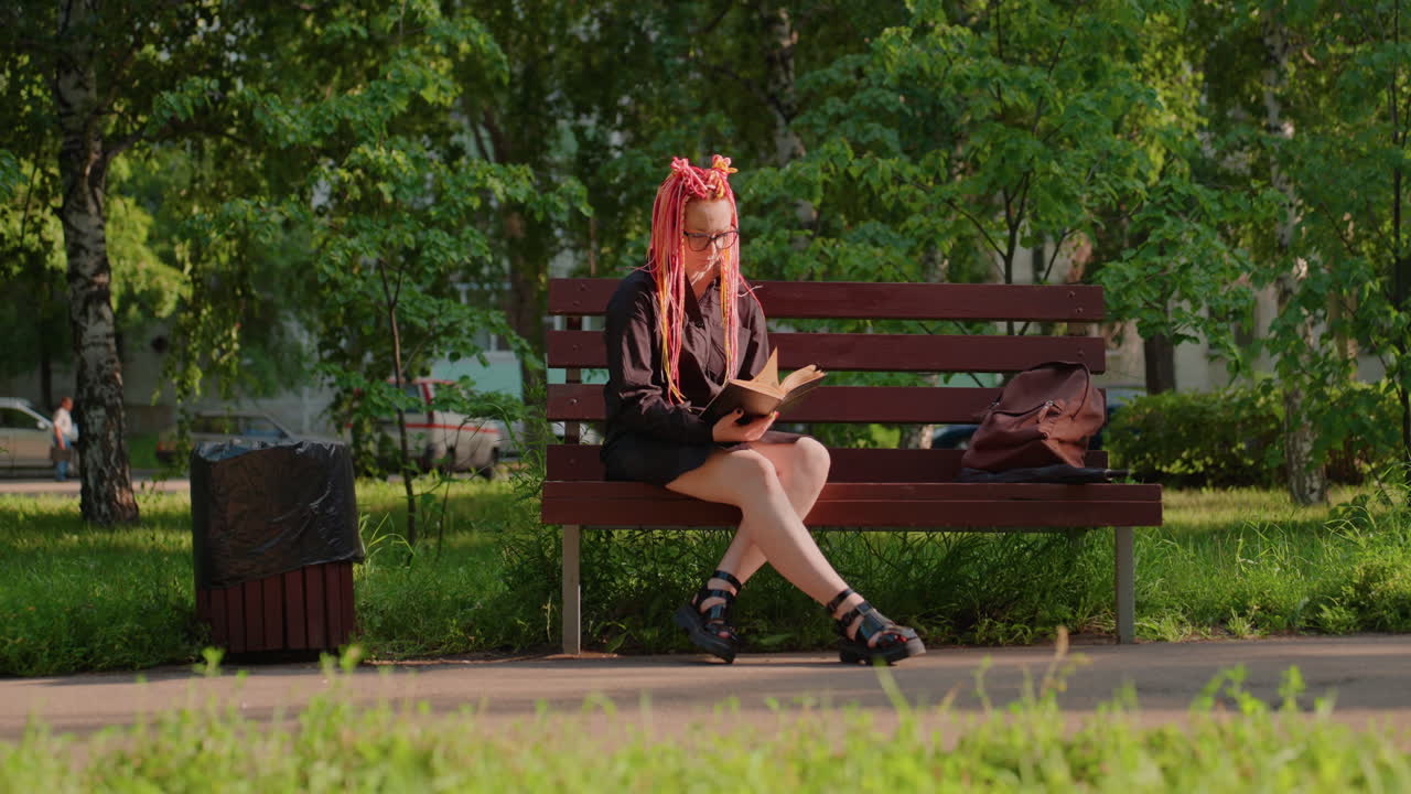 Mujer joven con pelo rosa leyendo un libro en un banco iluminado por el sol en un parque verde, coches y árboles a lo lejos de fondo, postura relajada, ropa informal y botas, auriculares alrededor del cuello, paneo cinematográfico lento