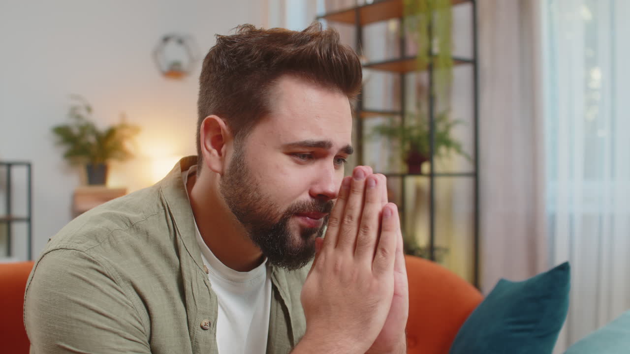 un joven caucásico tenso sentado en el sofá en casa doblando las palmas de las manos rezando pidiendo ayuda