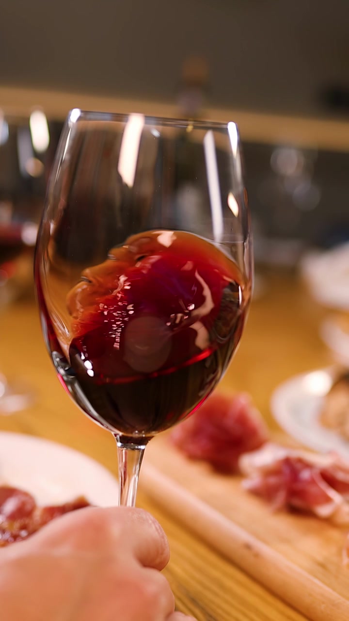 Una mano fa roteare vino rosso in un bicchiere su un tavolo con pizza e prosciutto