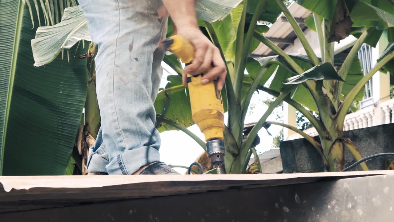 trabajador de la construcción que usa una herramienta de perforación para atornillar una hoja de zinc en una correa de ca que es parte de una estructura metálica en un sitio de construcción rural