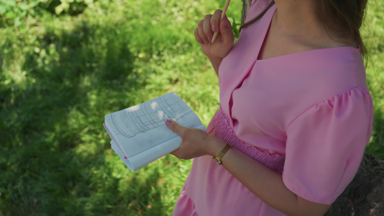 young caucasian woman journaling under tree, pink dress bathed in dappled sunlight, closeup hands holding pencil and textured notebook, leaning against trunk in grassy park, nostalgic summer mood