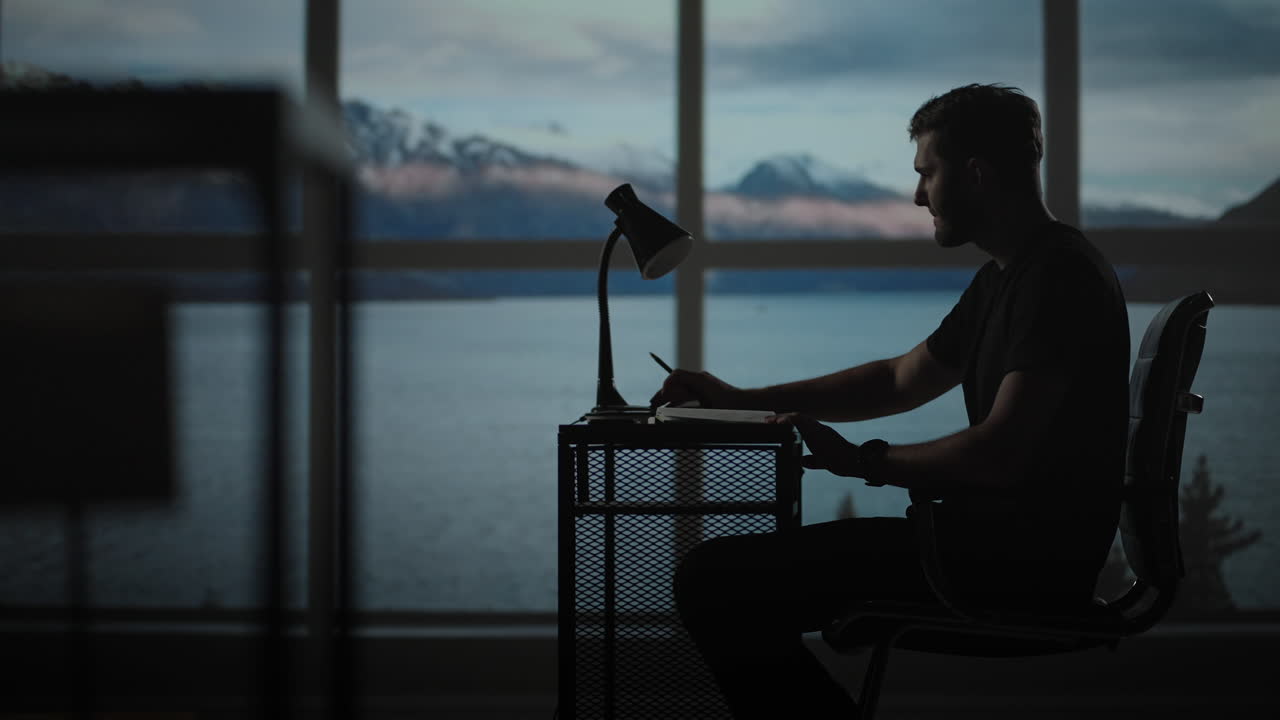 Serious concentrated young male student studying at home office sitting in cozy workplace at night under lamplight. A young man thinks creates a creative project and writes it down with a pen.