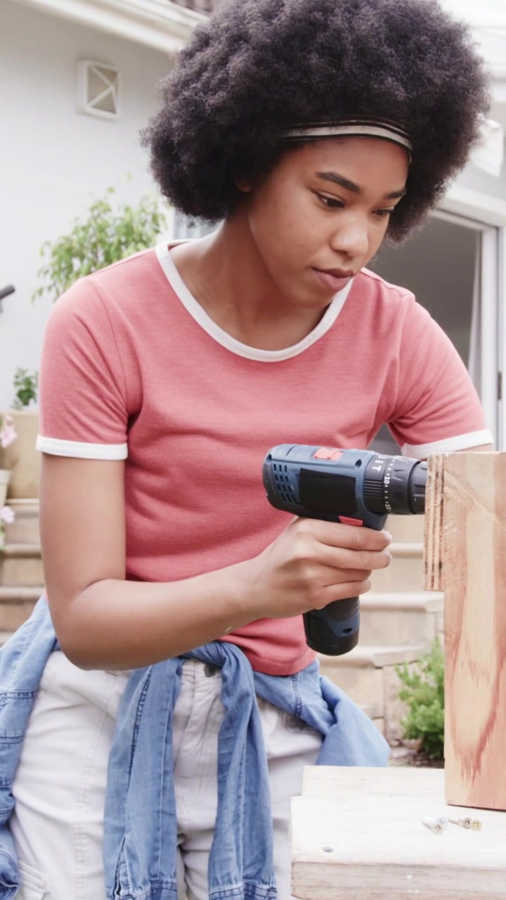 Video verticale: Donna con un trapano che fissa un pannello avvitando viti su un portico, con una camicia di jeans annodata.