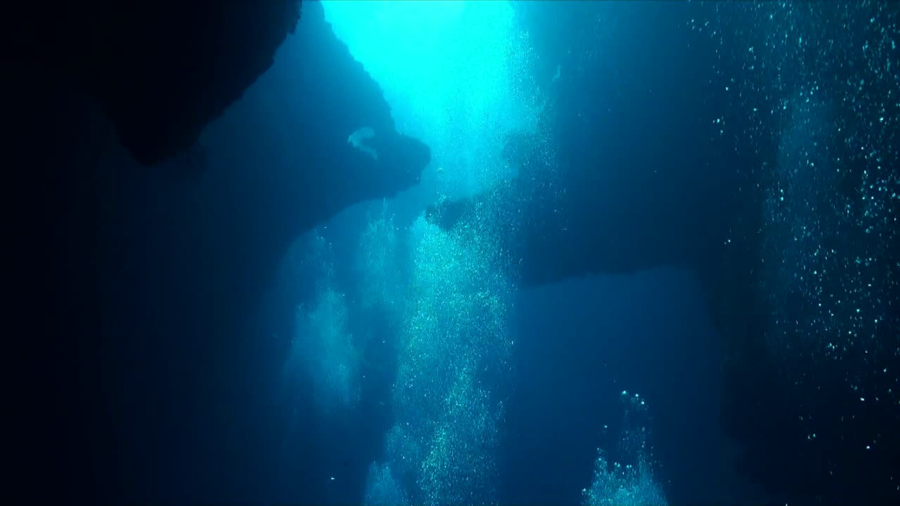Many air bubbles from scuba divers rising to the surface, wide angle shot with cave as backdrop