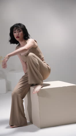 vertical video: model in stylish beige outfit posing confidently on cube in modern studio