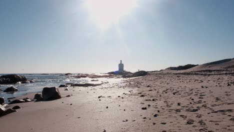 Weitwinkelaufnahme-Eines-Strandes-Mit-Kleinen-Felsen-Und-Einem-Leuchtturm-In-Der-Ferne