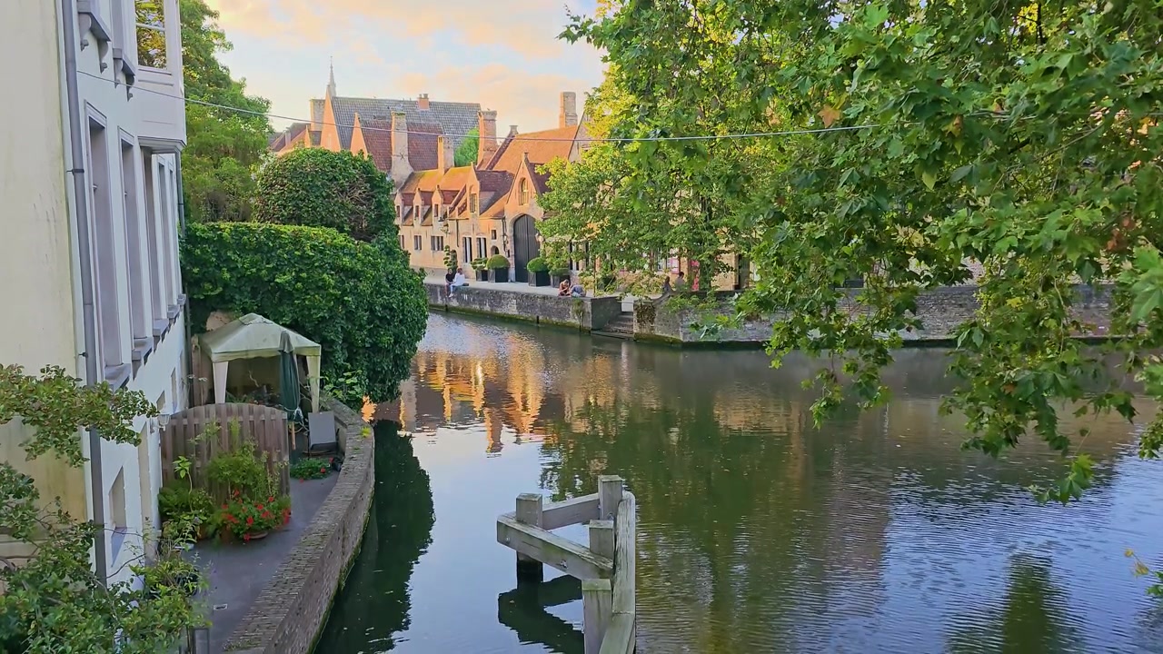 Suggestiva vista del canale con alberi verdi lussureggianti, case storiche sul lungomare, riflessi calmi e una tranquilla terrazza sull'acqua, creando una serena scena di viaggio europea al tramonto.