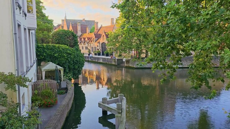 Suggestiva vista del canale con alberi verdi lussureggianti, case storiche sul lungomare, riflessi calmi e una tranquilla terrazza sull'acqua, creando una serena scena di viaggio europea al tramonto.