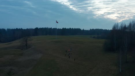aerial drone video flying over a field. flag fluttering in the wind, group of people walking across the field towards the flag. cloudy dark sky. 4k footage