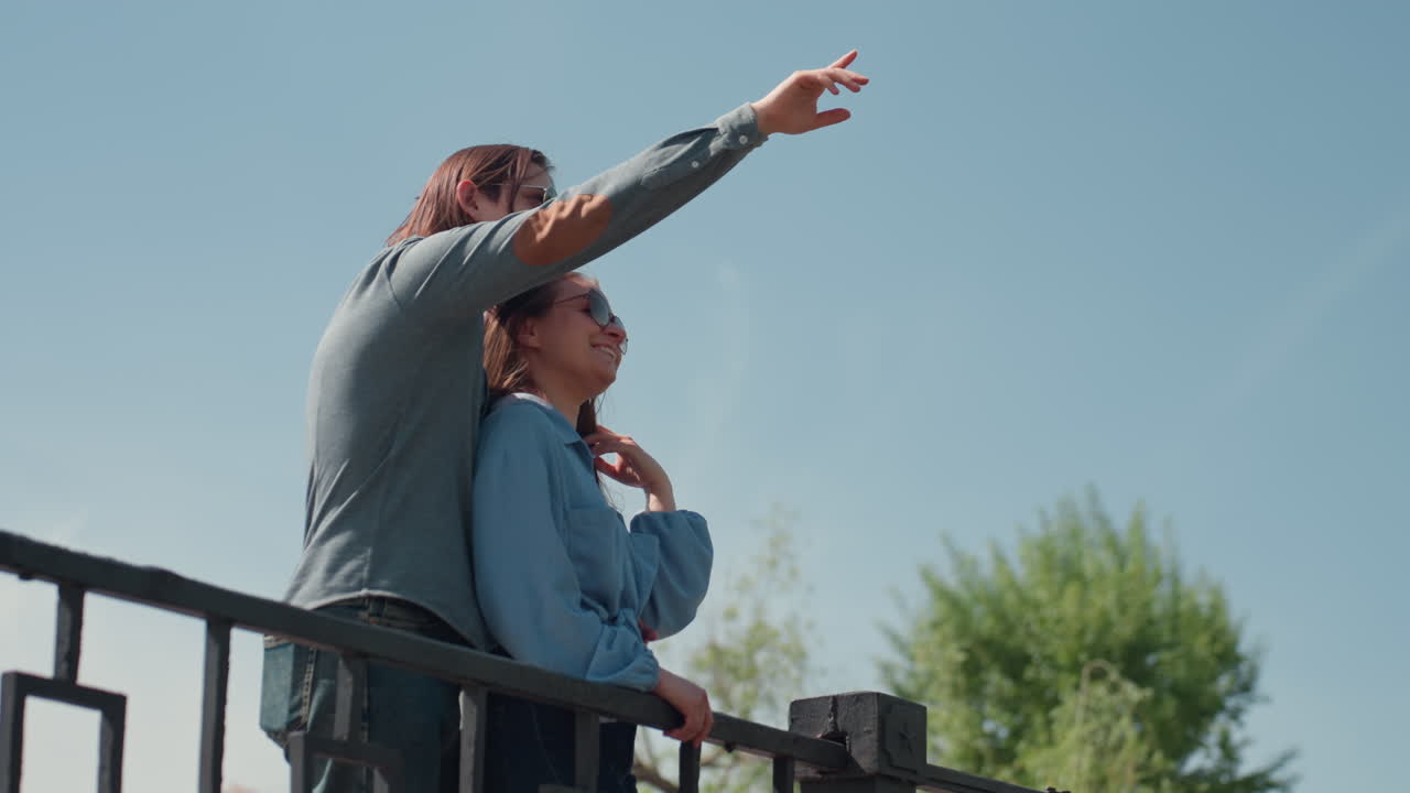 Белая пара на балконе показывает на ясное голубое небо, мужчина нежно обнимает женщину, указывая на далекий горизонт, повседневная джинсовая одежда, солнечный полдень, листва парка на заднем плане, атмосфера надежды