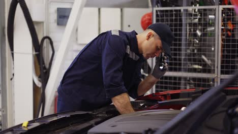 car service worker examinating engine under hood with flashlighter in hand, side view