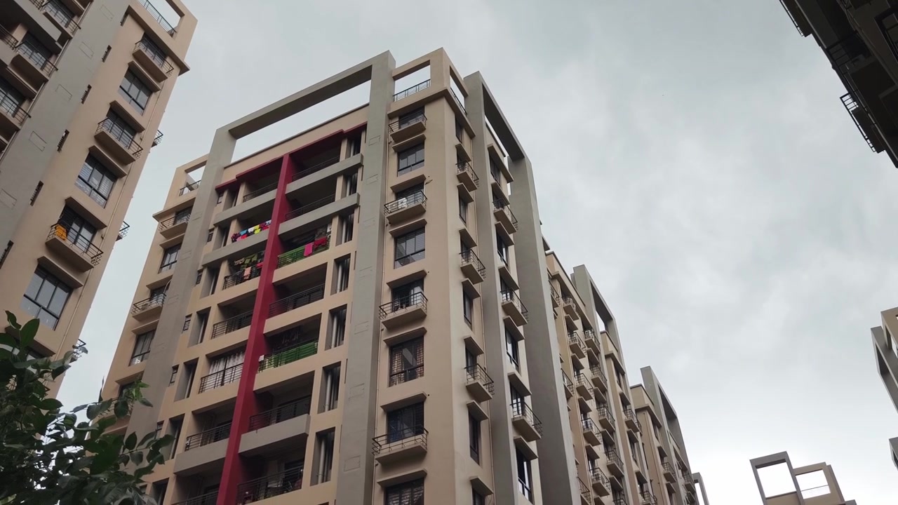 Plano panorâmico em ângulo baixo de complexos de apartamentos modernos e altos na Índia, com varandas e detalhes arquitetónicos vermelhos sob um céu nublado.