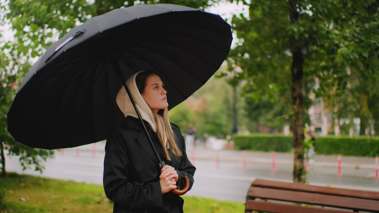 Young woman in hooded coat holding umbrella walking near road on rainy day, looking up with thoughtful expression, blurred background with trees and pedestrians, calm urban autumn lifestyle moment