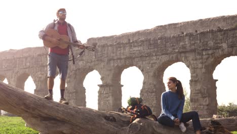 happy young couple backpackers tourists on a log trunk playing guitar singing in front of ancient roman aqueduct ruins in romantic parco degli acquedotti park in rome at sunrise slow motion