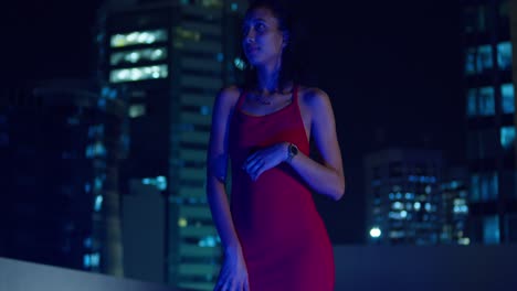 una joven con un vestido rojo está en un techo por la noche, los edificios de la ciudad se vislumbran en el fondo