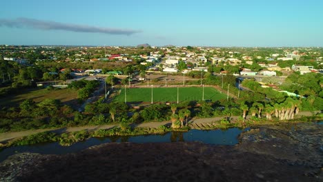 Aerial-overview-of-green-grass-soccer,-football-field-in-Caribbean-neighborhood,-next-to-toxic-asphalt-lake-from-refinery