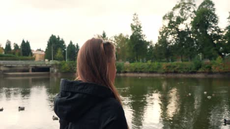 Young-beautiful-girl-with-long-hair-looks-on-the-shore-of-the-pond,-turns-to-the-other-side-and-walks-away