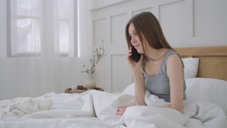 young caucasian woman sitting on the bed in the morning feels bad. a young woman is making a smartphone call to her doctor.