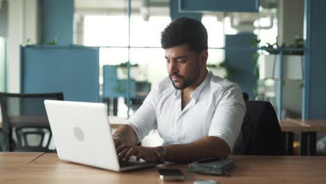 stylish-young-man-of-Arab-descent-sitting-in-a-modern-business-center-office-with-a-laptop,-working-furiously,-upset-by-failure,-closing-the-laptop